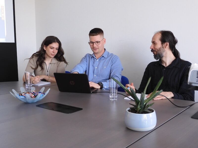 Drei Personen arbeiten gemeinsam an einem Laptop in einem modernen Büro mit Besprechung.