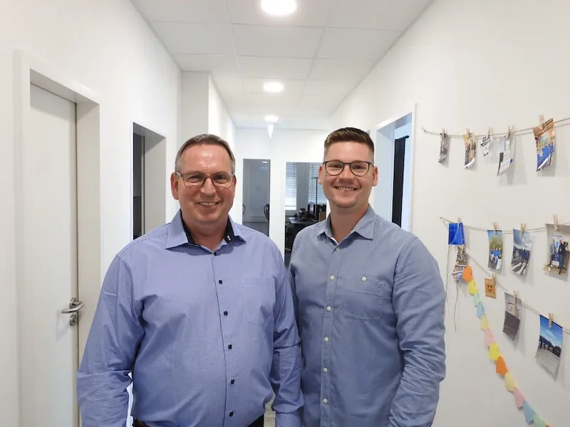 Zwei lächelnde Männer im Büroflur mit Fotos an der Wand, beide in blauen Hemden.