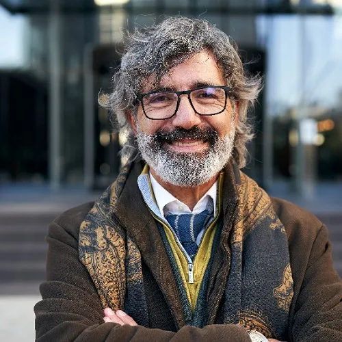 Ein älterer Mann mit Brille und Bart lächelt vor einem modernen Bürogebäude.