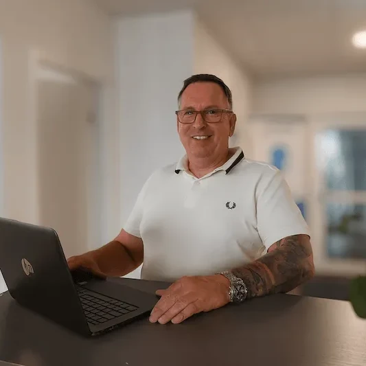 Mann mit Brille arbeitet lächelnd an einem Laptop im modernen Büro mit Tageslicht.