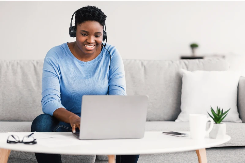 Frau mit Headset arbeitet im Homeoffice am Laptop auf dem Sofa, Videokonferenz, Remote Work.